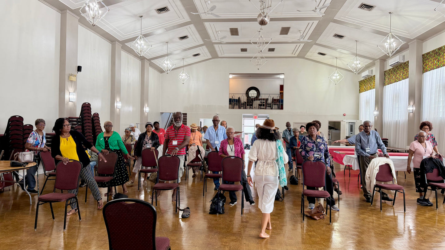 A large indoor omax3m Maxine Yoga space with a high coffered ceiling, chandeliers, wood floors, and many people standing and sitting around tables and folded chairs. The scene appears to be a banquet hall or community event rather than a clean product listing photo.