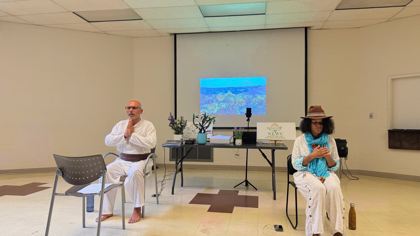 Maxine and client seated on chairs in a room with a projector screen, table, and presentation setup. The person on the left wears a white robe and bow tie, and the person on the right wears a white outfit with a turquoise scarf and brown hat.