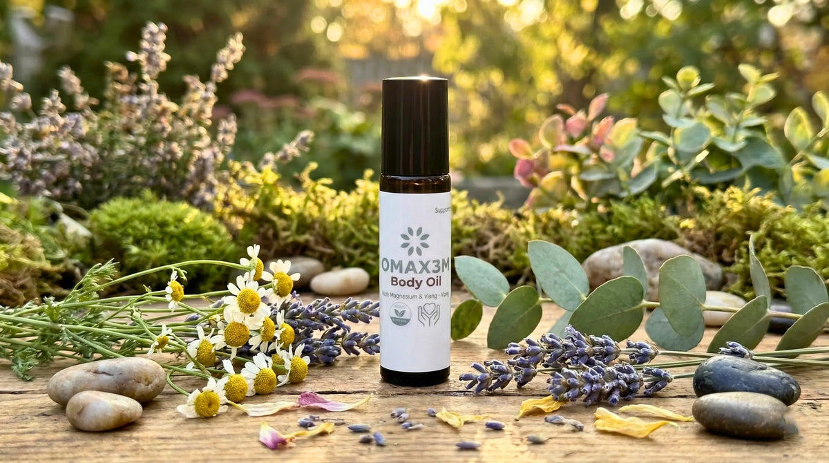 A bottle of OMAX3N Body Oil stands on a wooden surface surrounded by natural props including eucalyptus leaves, lavender sprigs, chamomile flowers, and smooth stones, with a blurred garden background.