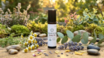 A bottle of OMAX3N Body Oil stands on a wooden surface surrounded by natural props including eucalyptus leaves, lavender sprigs, chamomile flowers, and smooth stones, with a blurred garden background.