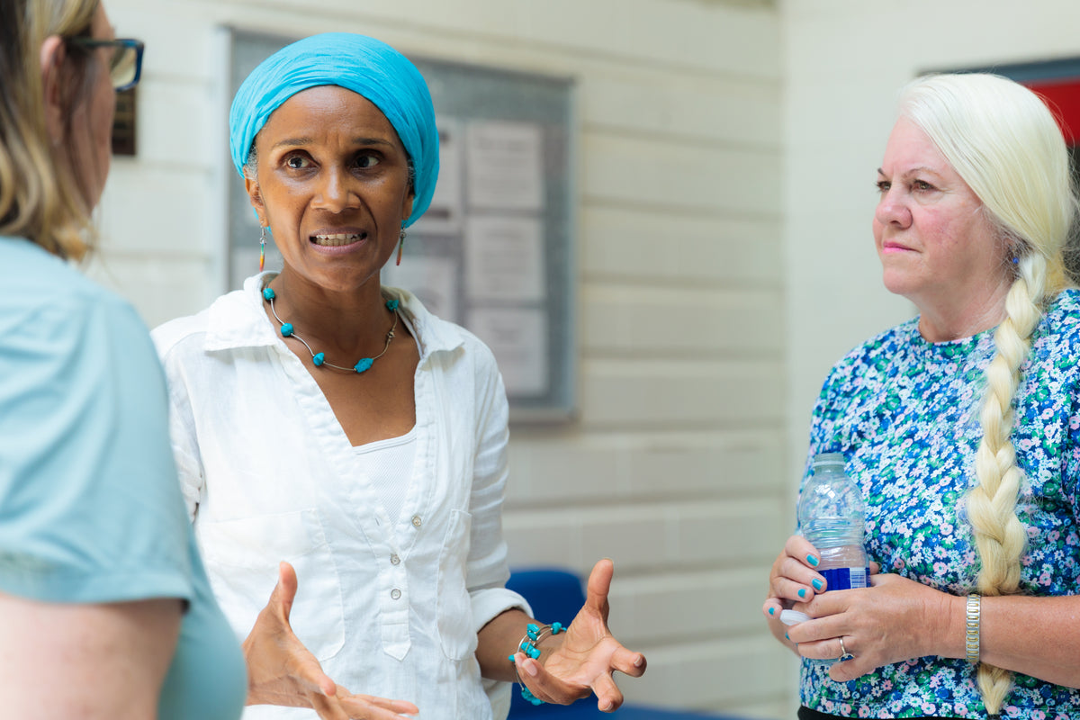 Maxine in a white shirt and bright blue headwrap is speaking animatedly with two other women in a light-colored indoor setting. The central woman has turquoise jewelry and a matching turquoise headwrap, while the woman on the right wears a blue and white patterned top and holds a water bottle.