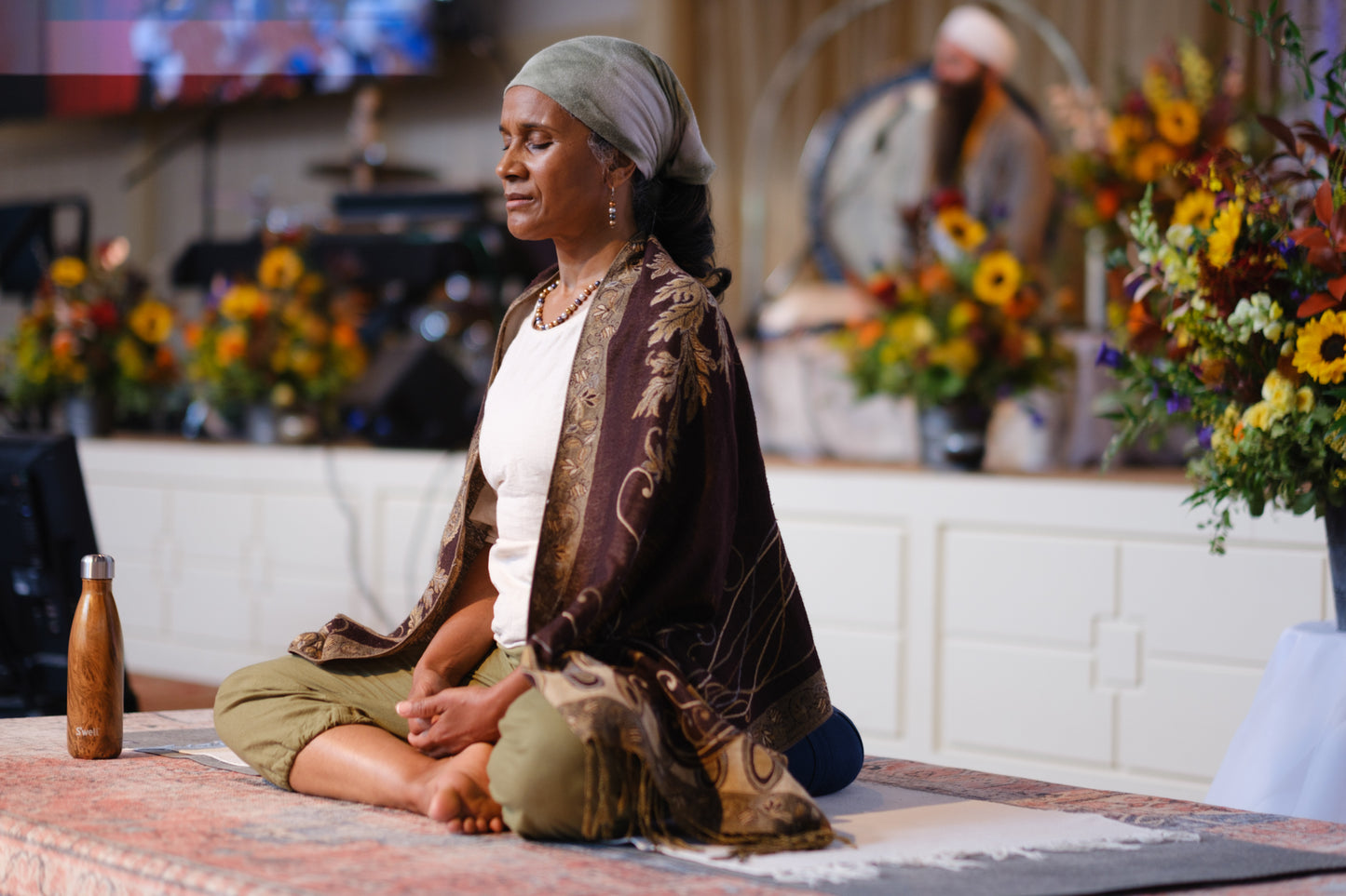 Maxine sits cross-legged on a patterned rug in a decorated indoor space, wearing a white top, a brown patterned shawl, and olive green pants. A small wooden bottle is placed beside her.