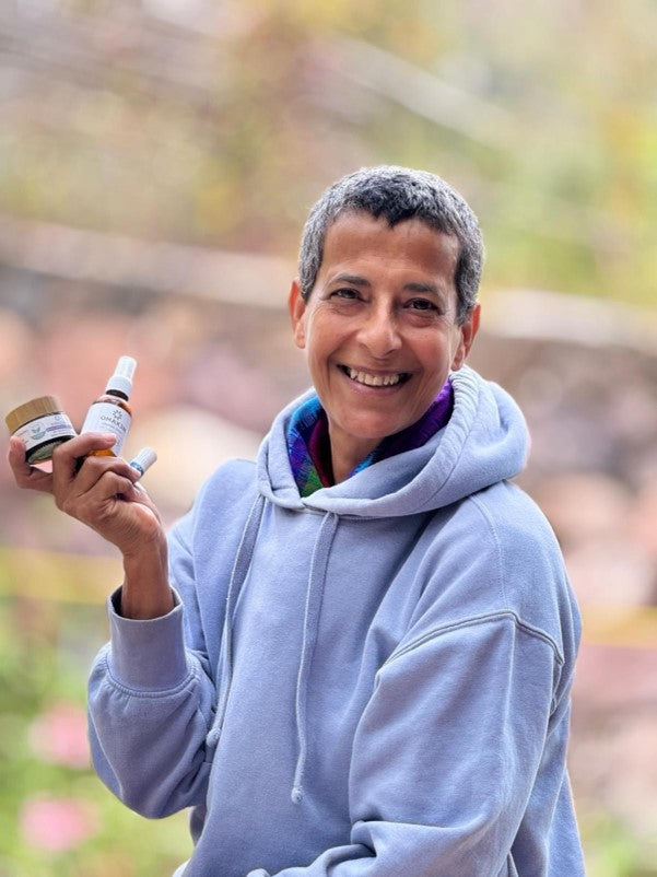 Claudia smiling and holding OMAX3M natural skincare products, including magnesium cream body butter and calming spray for wellness.